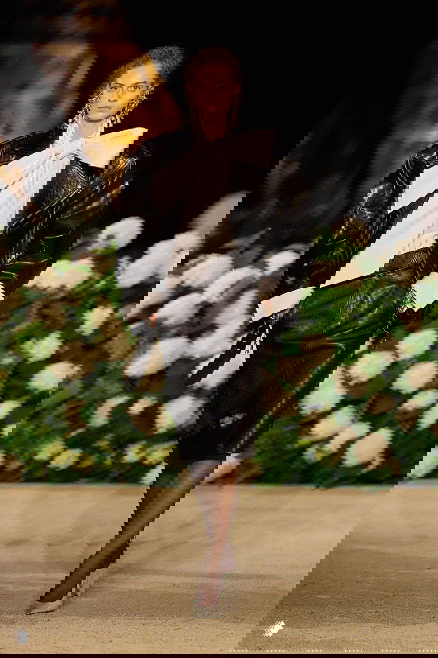 Модель в чёрном кожаном жакете и юбке Saint Laurent с большим белым бантом; дефиле на подиуме на фоне белых цветов и тёплого освещения.