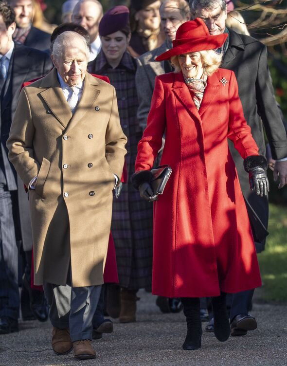 Двое людей идут рядом: женщина в длинном алом пальто и мужчина в коричневом пальто; вокруг — другие участники праздничной ходьбы у церкви.