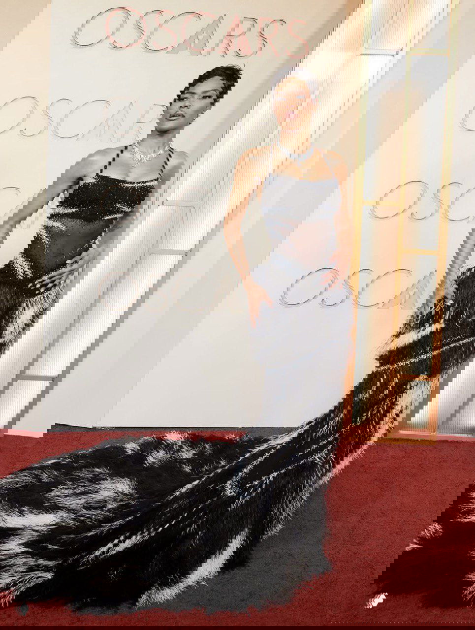 Женщина в эффектном тёмно-черном платье с фактурным шлейфом и блеском на фоне декора Оскара 2026, позирует для фотографов.