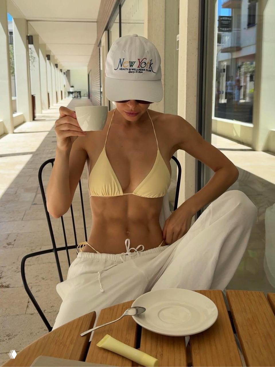 Модель в бикини и кепке сидит за столом на улице, видно выраженную прокачку пресса и тонкую талию в летнем окружении кафе.