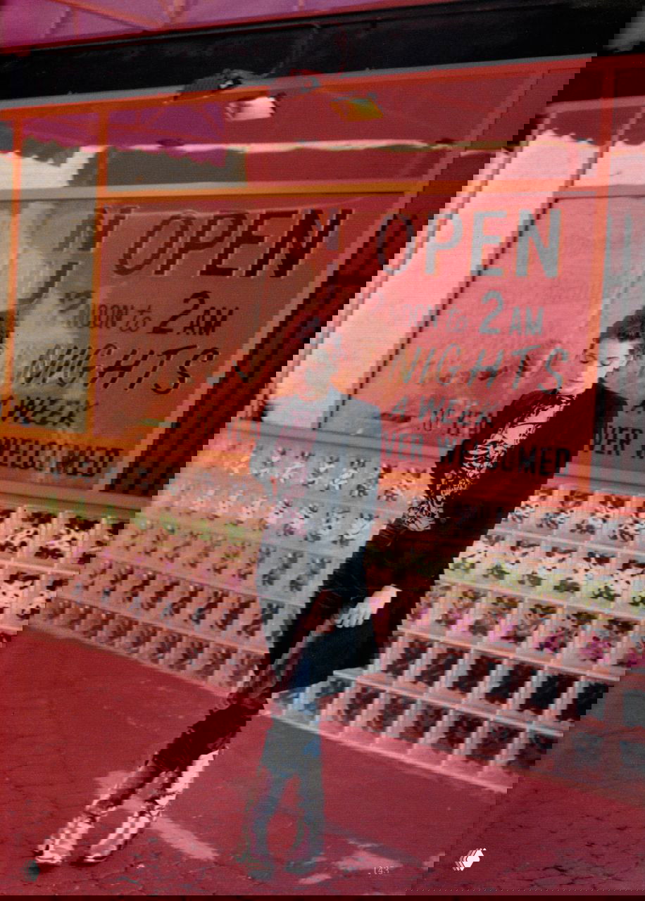 Уличный кадр с вывеской и неоновым оттенком: модель в длинном пиджаке и блестящих сапогах идёт вдоль витрины, городской вечер и контрастные цвета подчёркивают образ.