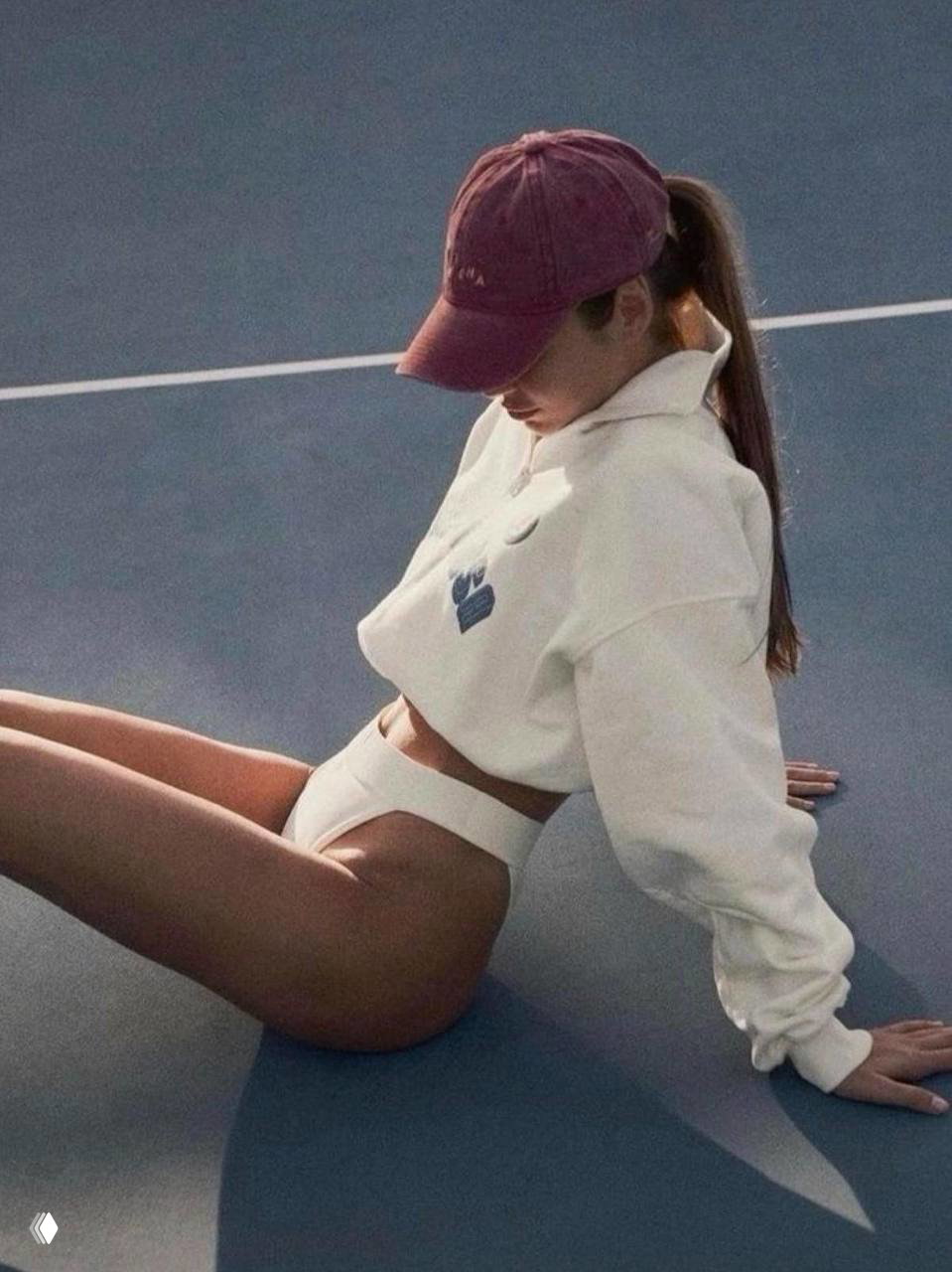 Девушка в свитшоте и плавках сидит на корте, поза демонстрирует растяжение и расслабление после нагрузки на ноги и корпус.