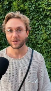 Мужчина в светлом свитере говорит в микрофон на фоне густой зелёной листвы; кадр интервью на мероприятии, лицо выражает живую эмоциональную реакцию.