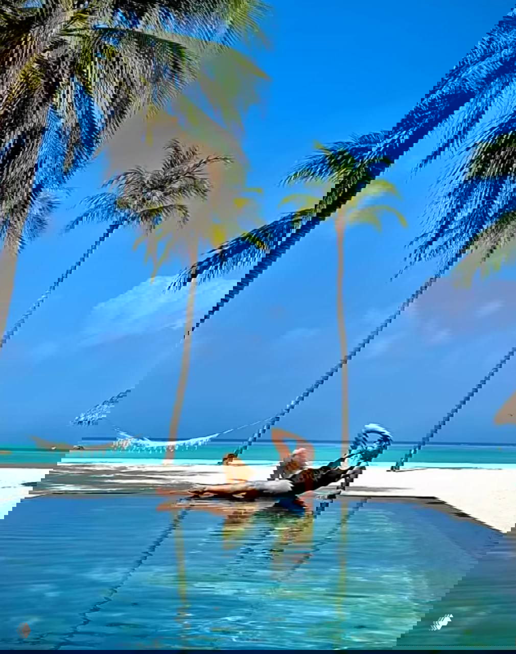 Пляжный пейзаж Мальдив: бассейн у белого песка, пальмы и женщина в купальнике на берегу — яркий тропический отдых.