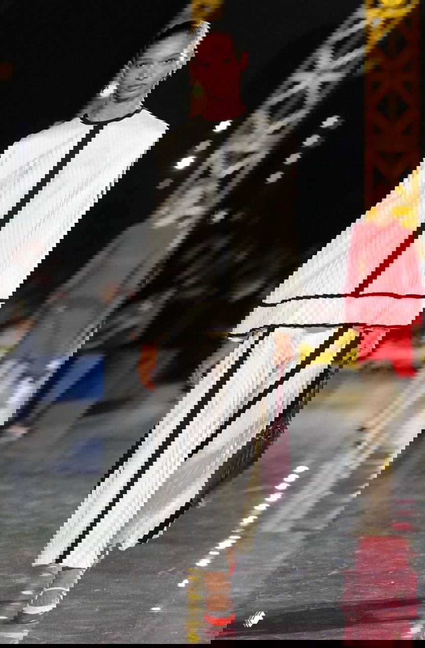 Модель в белом комплекте с чёрными полосами выходит на подиум Chanel; образ сочетает строгие линии и плиссированные брюки в контексте индустриального декора.