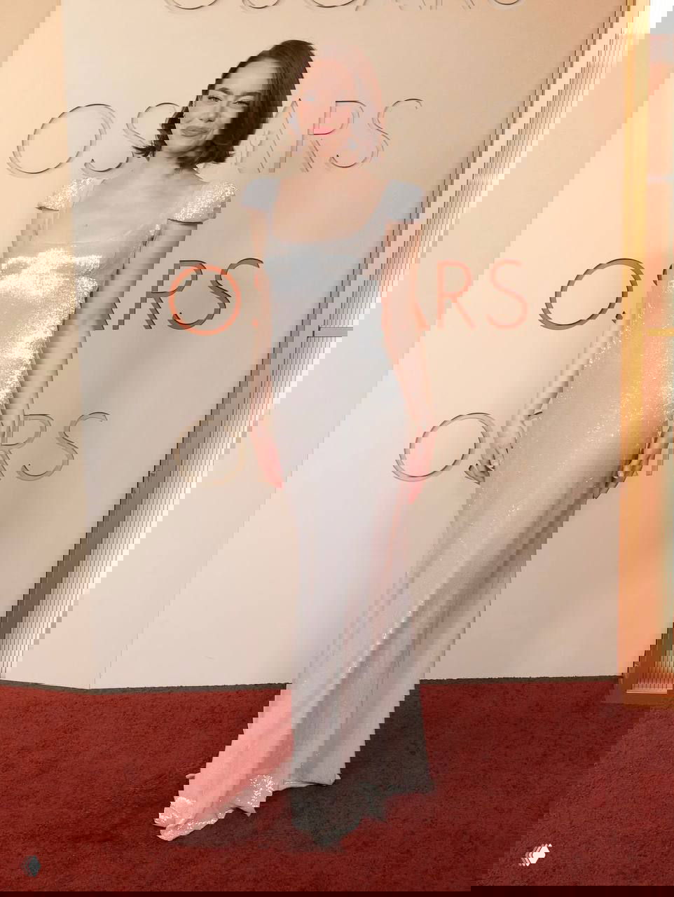 Женщина в серебристом струящемся платье в полный рост на фоне логотипа «Oscars» — лаконичный и элегантный образ на красной дорожке 2026.