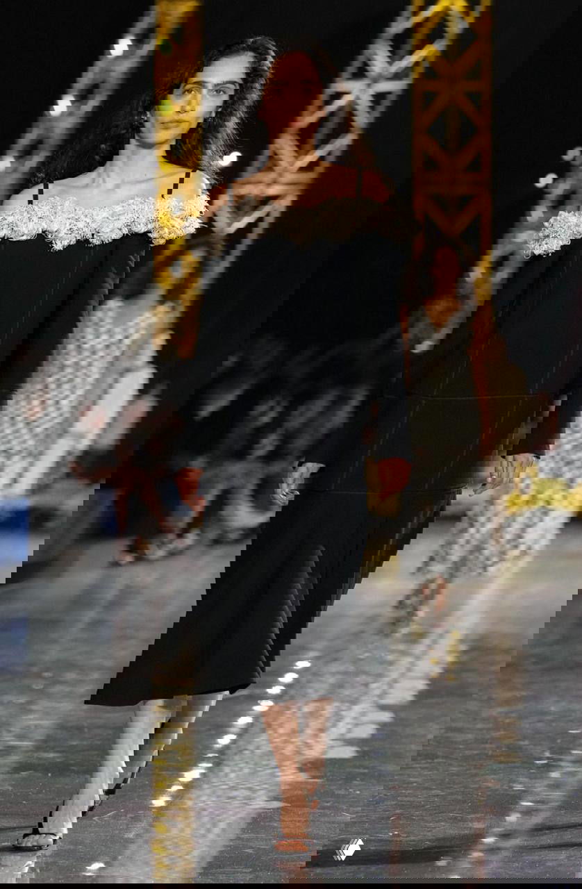 Модель в черном платье с открытыми плечами и цветочными брошь‑элементами проходит показ Chanel; акцент на классическую элегантность и декор плеч.