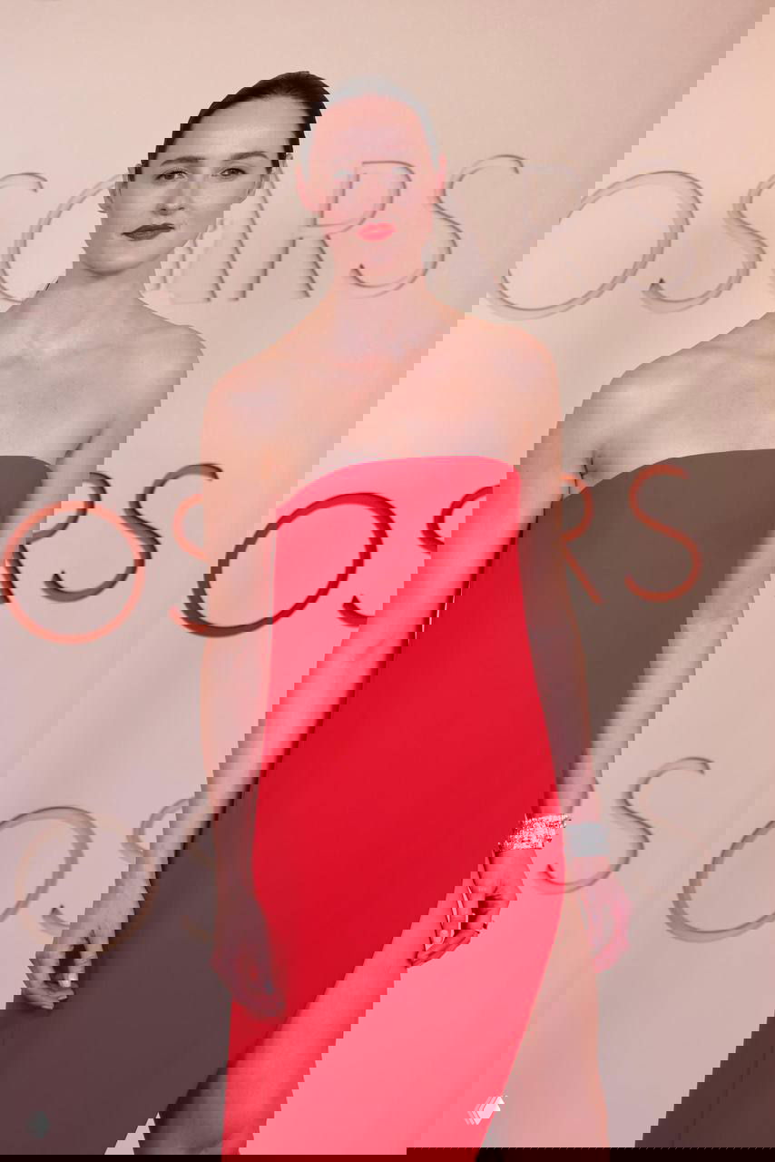 Женщина в ярком красном платье без бретелей с высоким разрезом на фоне фотозоны Оскара 2026, портрет в полный рост.