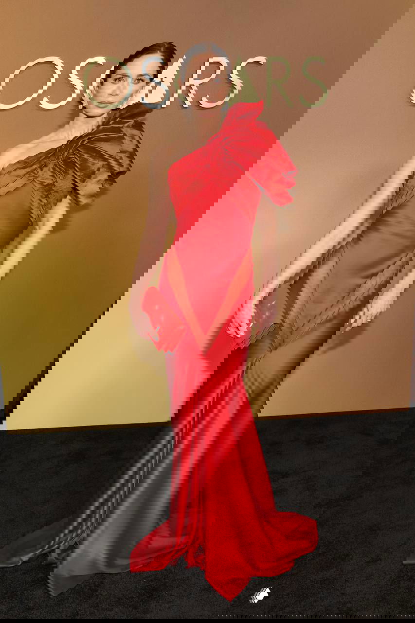 Женщина в приталенном ярко‑красном платье с крупным декоративным элементом на плече и маленьким клатчем позирует на красной дорожке у золотого баннера церемонии.