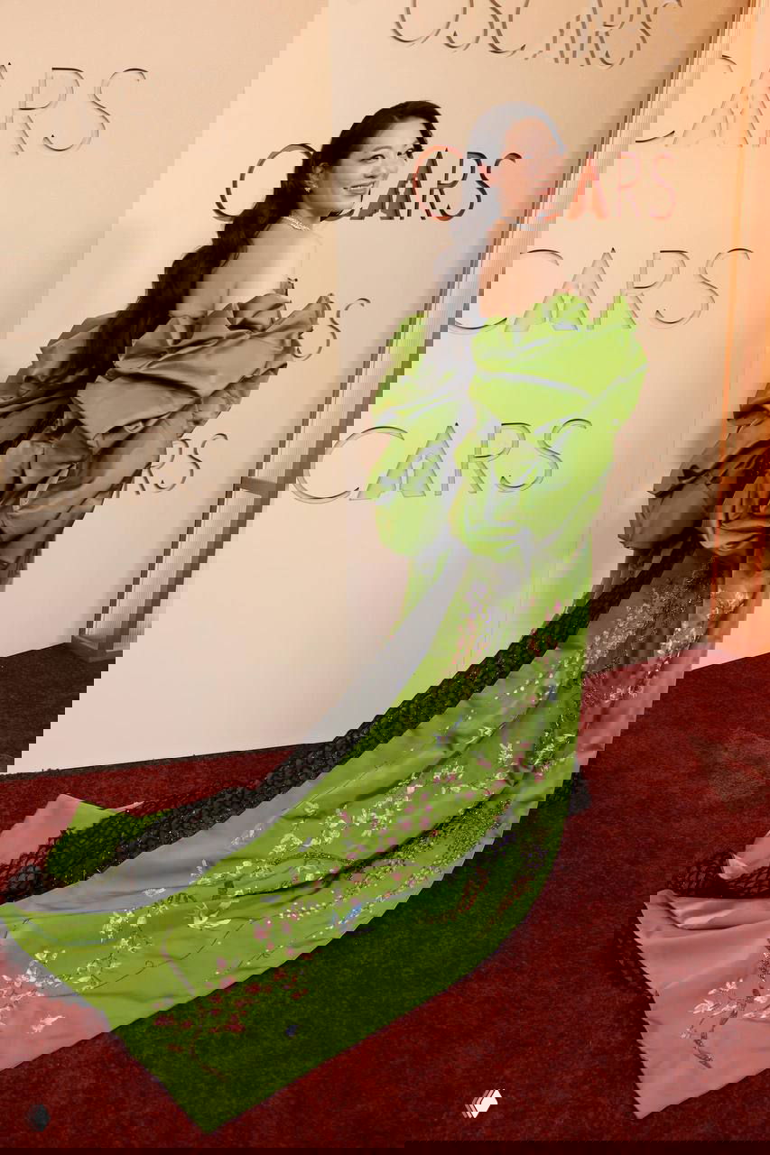 Женщина в зелёном платье с пышным шлейфом и декоративной вышивкой, позирует на красной дорожке Оскара 2026, вид со спины в три четверти.