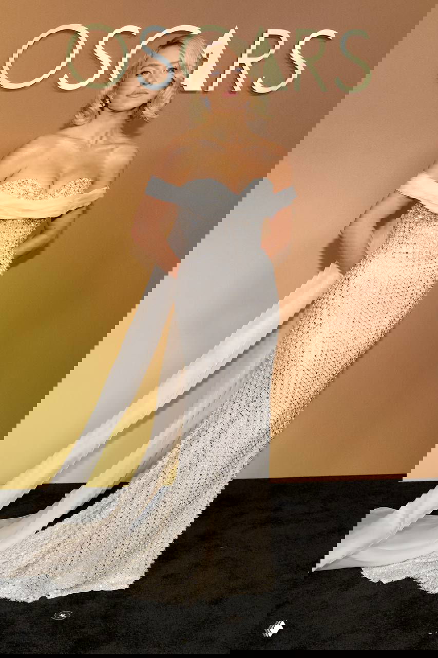 Женщина в сверкающем серебристом платье с открытыми плечами и драпировкой стоит у золотого фона на красной дорожке, утончённый вечерний образ с блеском тканей.