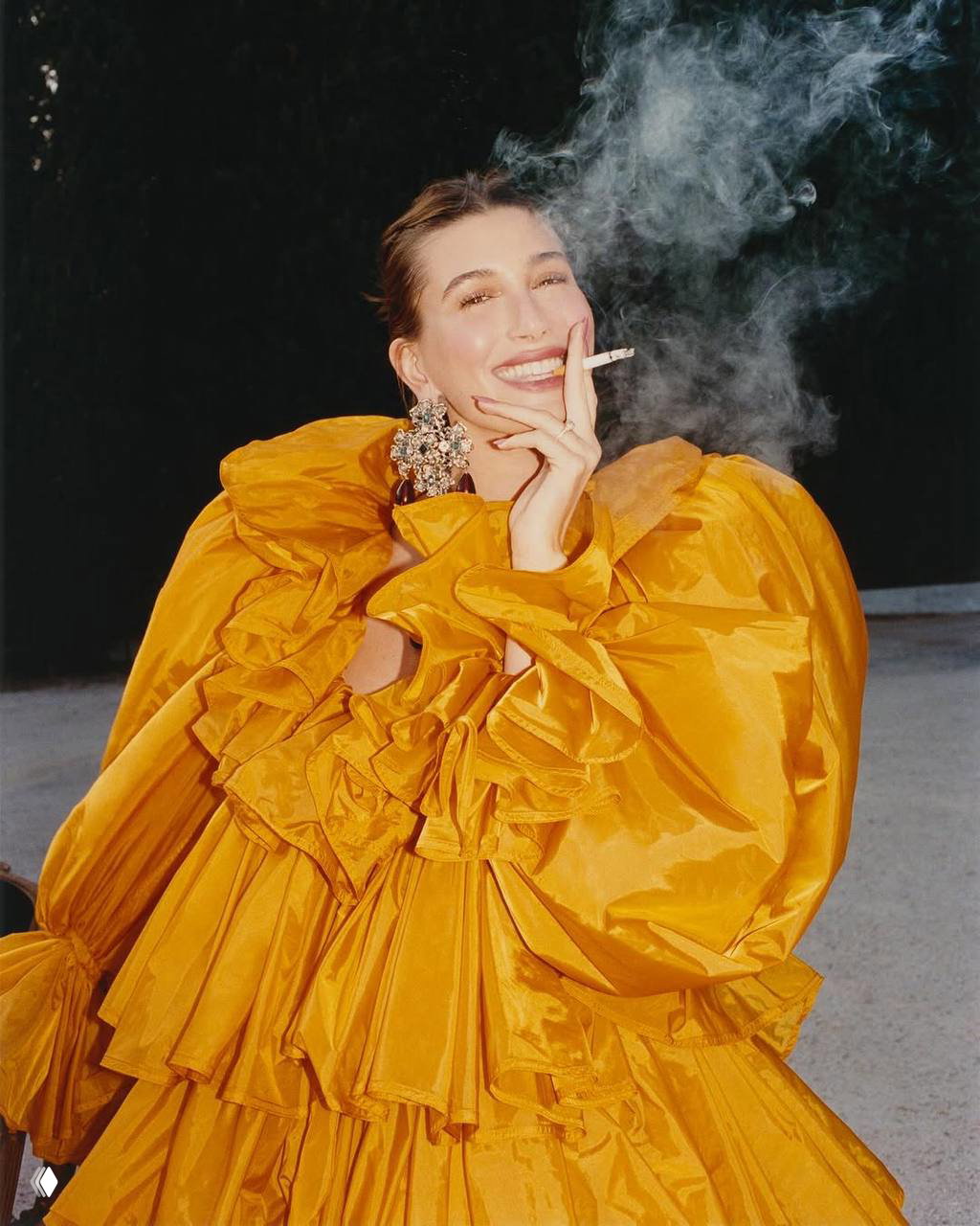Крупный план модели в пышном жёлтом платье: она держит сигарету, клубы дыма вокруг, выразительная улыбка и крупная брошка.