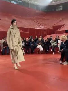 Модель в бежевом тренче дефилирует по насыщенно‑красному подиуму показа Lacoste на Парижской неделе моды, зрители в первом ряду следят за показом.