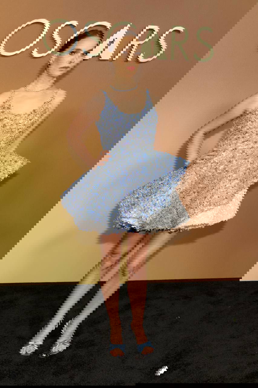 Женщина в коротком голубовато‑сером платье с пышной текстурной юбкой и сияющей отделкой на фоне золотого баннера церемонии, игривый вечерний силуэт.