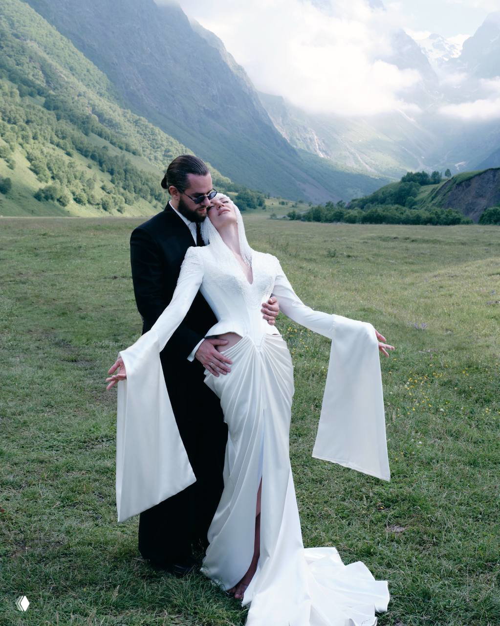 Молодожёны обнимаются на цветущем лугу в горах Северной Осетии: невеста в струящемся платье, жених в чёрном костюме — кадр с живописным пейзажем.
