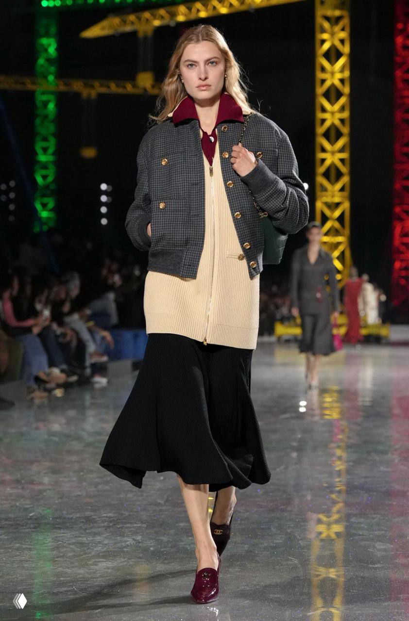 Модель в тёплом пуховом или пальтовом образе и расклешенной юбке идет по подиуму Chanel; в кадре видны зрители и индустриальные конструкции шоу.