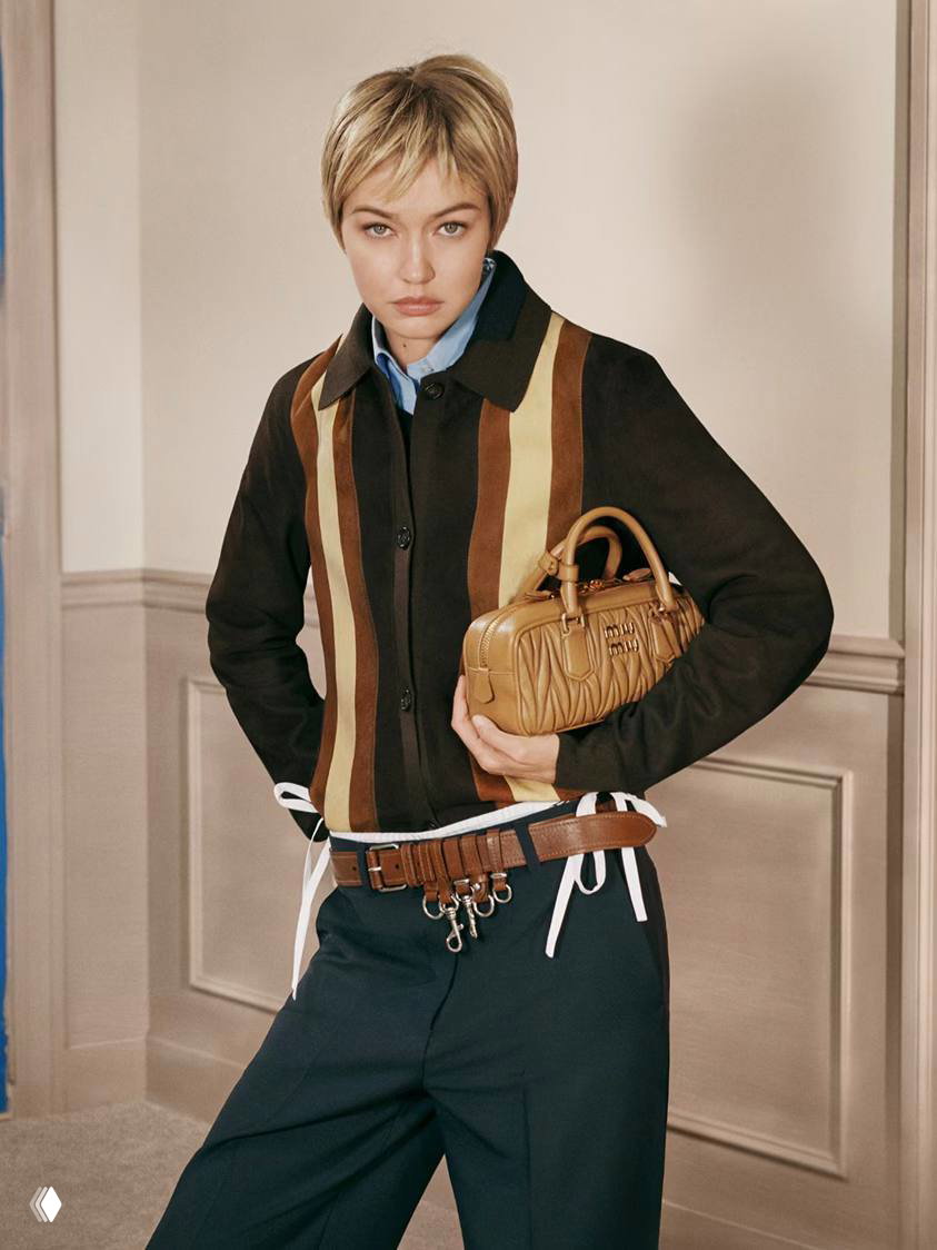 Модель в коротком парике в полной ростовой позе: коричневая куртка, сумка-баул и аксессуары на поясе в стилистике кампейна Miu Miu.
