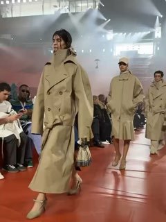 Группа моделей в бежевых плащах и тренчах шагает по красному покрытию подиума на демонстрации Lacoste во время Парижской недели моды, фотографы и гости по бокам.