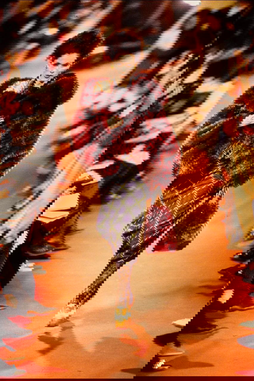 Модель в компактной бордовой пушистой шубе и клетчатой юбке идёт по подиуму красного цвета на показе Vivienne Westwood 1993.