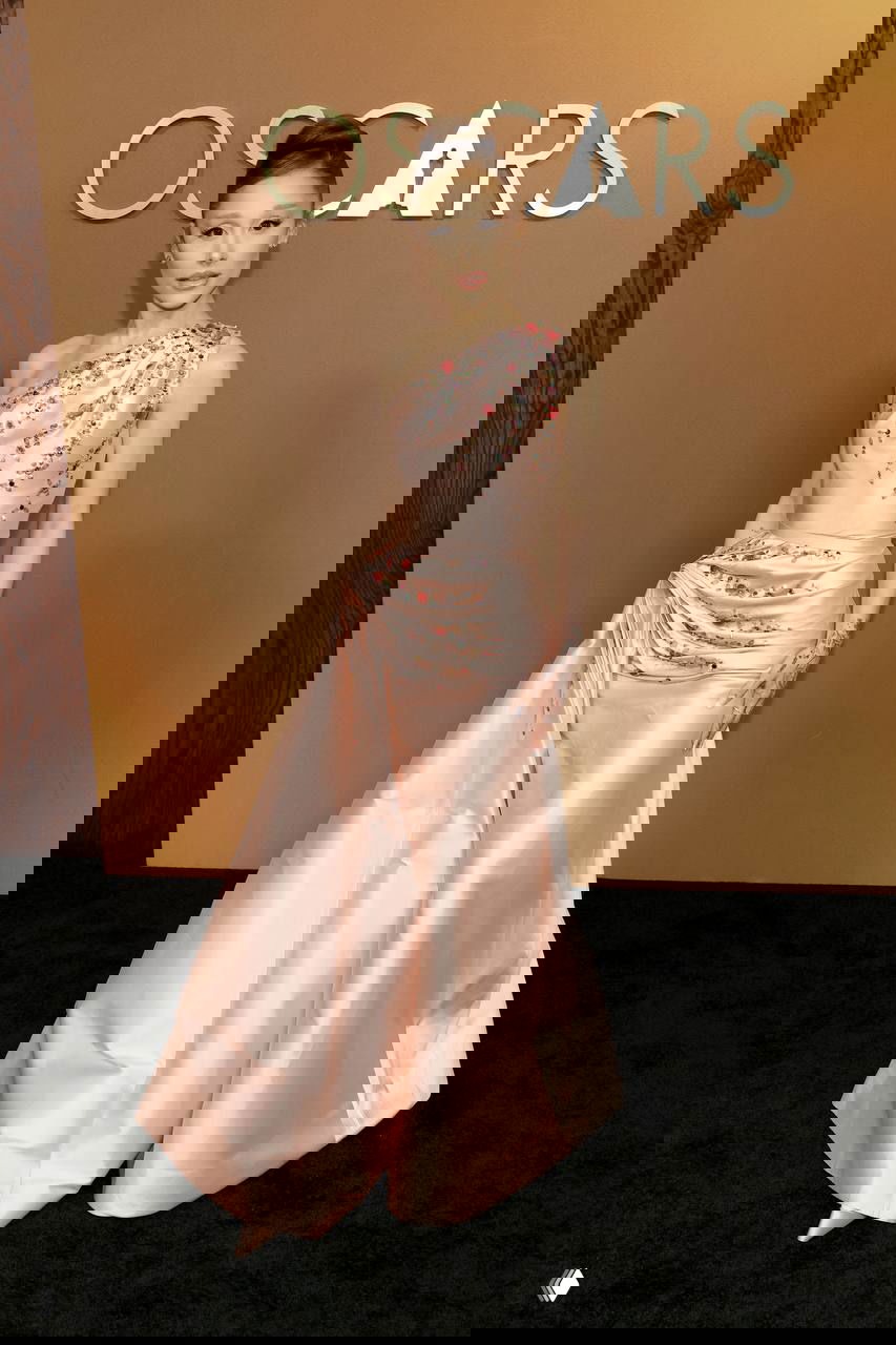 Женщина в розовом атласном платье с декоративной драпировкой и расшитыми деталями стоит у золотого баннера Oscars, изысканный вечерний образ с текстурой ткани.