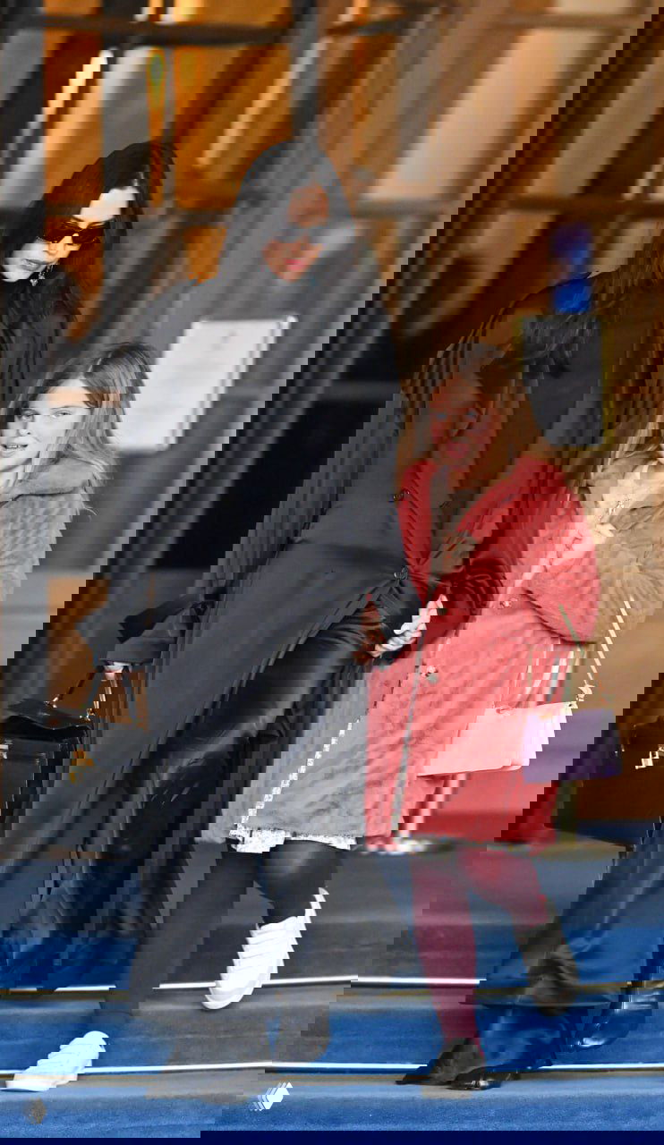 Фото: женщина в длинном чёрном атласном пальто выходит из гостиницы вместе с девочкой в розовой шубке, городской вход и ступени.