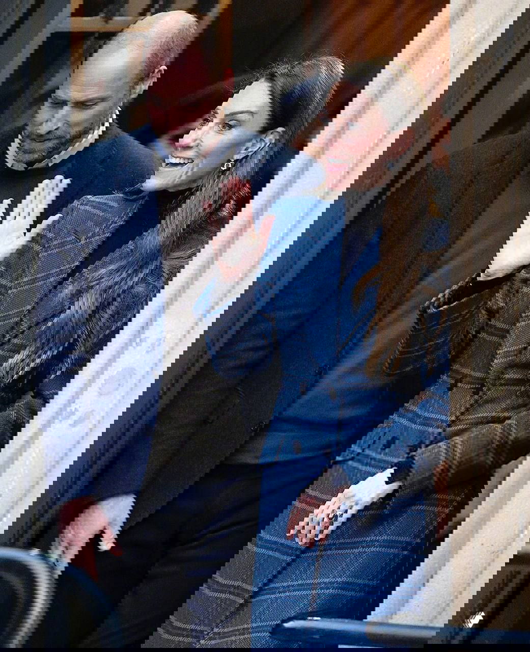 Улыбчивый кадр: пара выходит из здания, женщина в пальто в клетку машет рукой, дневной городской фон, официально-повседневный образ.