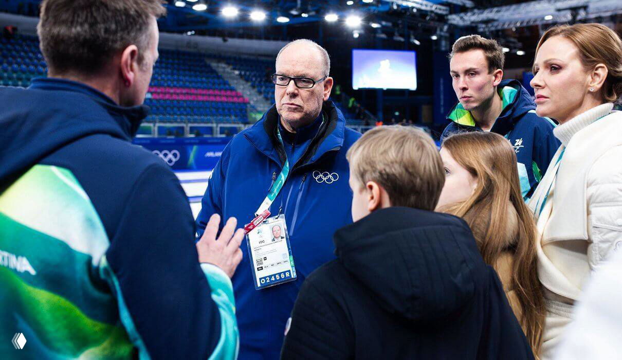 Князь Альбер II и сопровождающие представители обсуждают соревнования с членами команд, на переднем плане видно детей и княгиню Шарлен в светлом пальто.