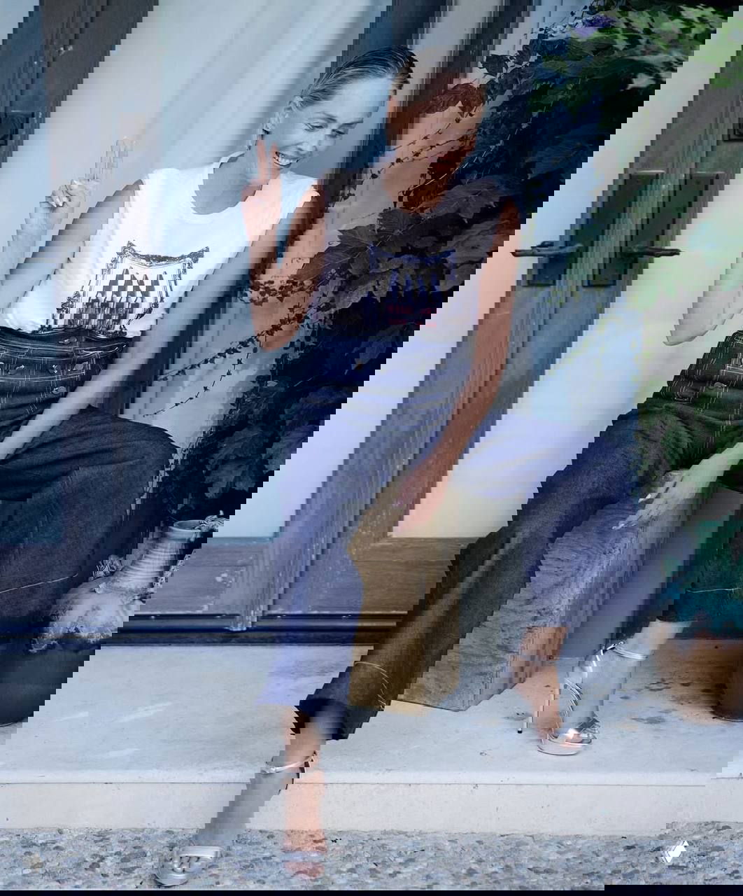 Женщина в белой майке и тёмных широких джинсах сидит на деревянном кубе у дверей, улыбается и показывает жест 'V' — портретный кадр.