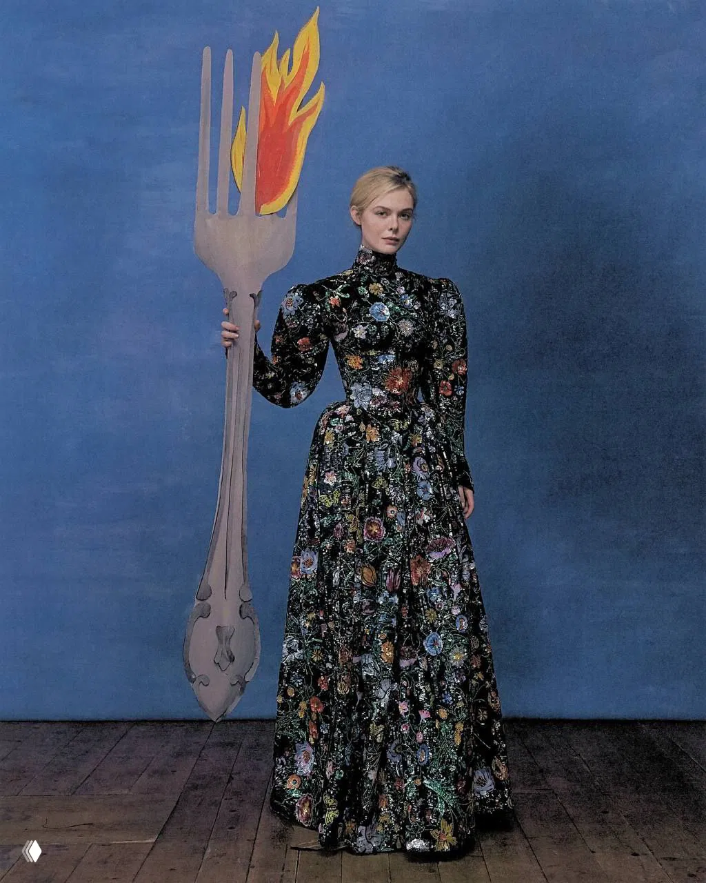 Полный рост Эль Фаннинг в длинном платье с цветочным узором держит крупную декоративную вилку с огнём — театральная постановка.