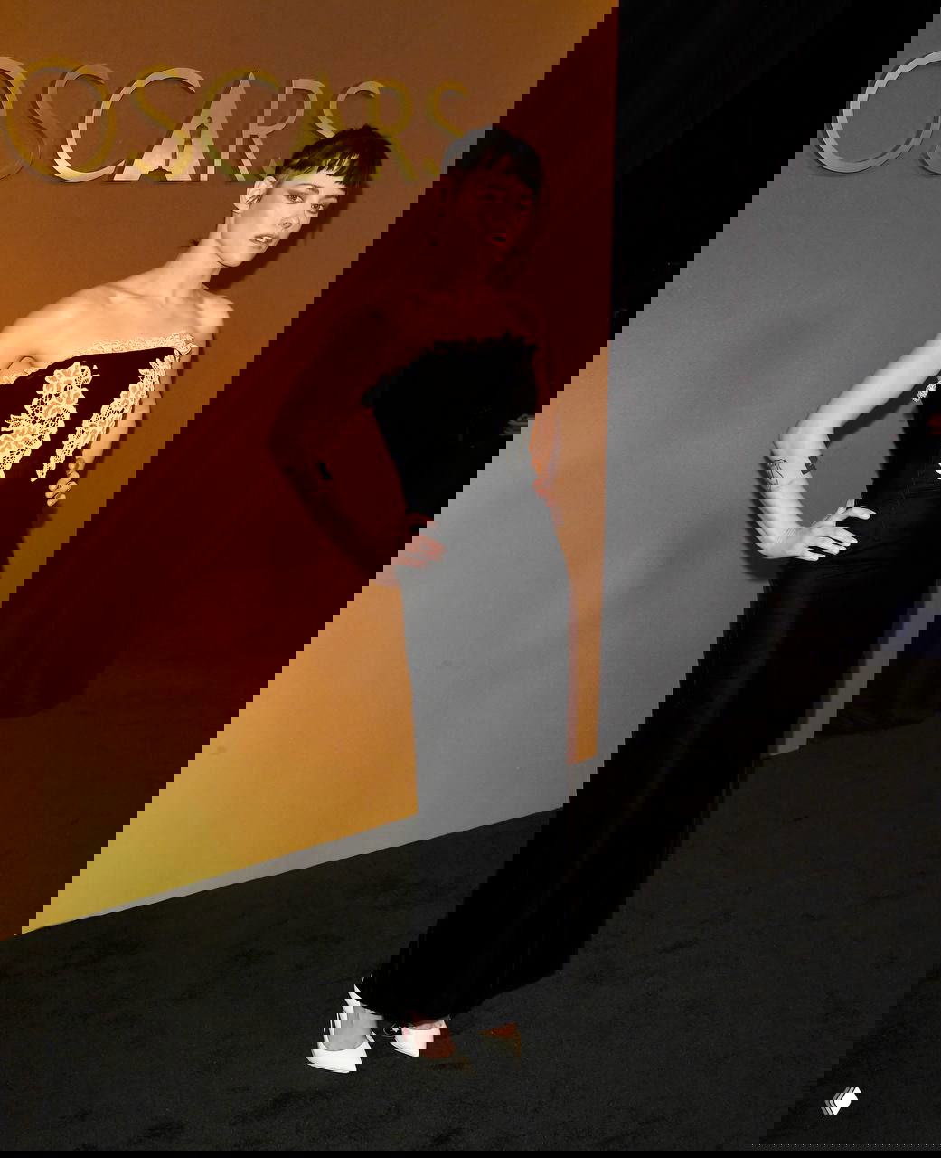 Женщина в чёрном платье‑футляре с кружевной аппликацией и короткой причёской позирует у золотого баннера Oscars на красной дорожке, элегантный и смелый образ.