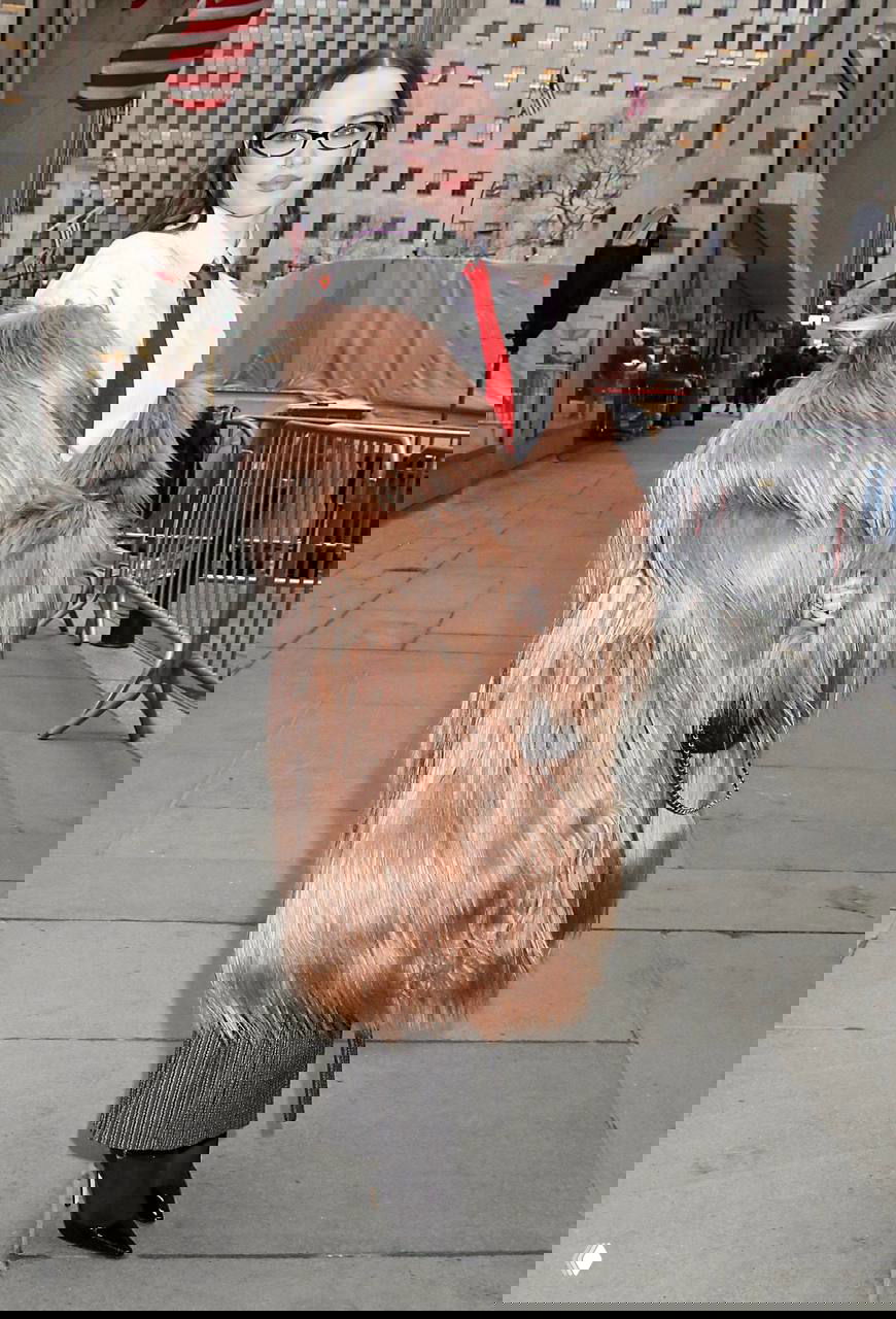 Дав Камерон идёт по тротуару в образе с ярко‑красным галстуком, светло‑коричневой объёмной шубой, чёрной сумкой на цепочке и лодочками.