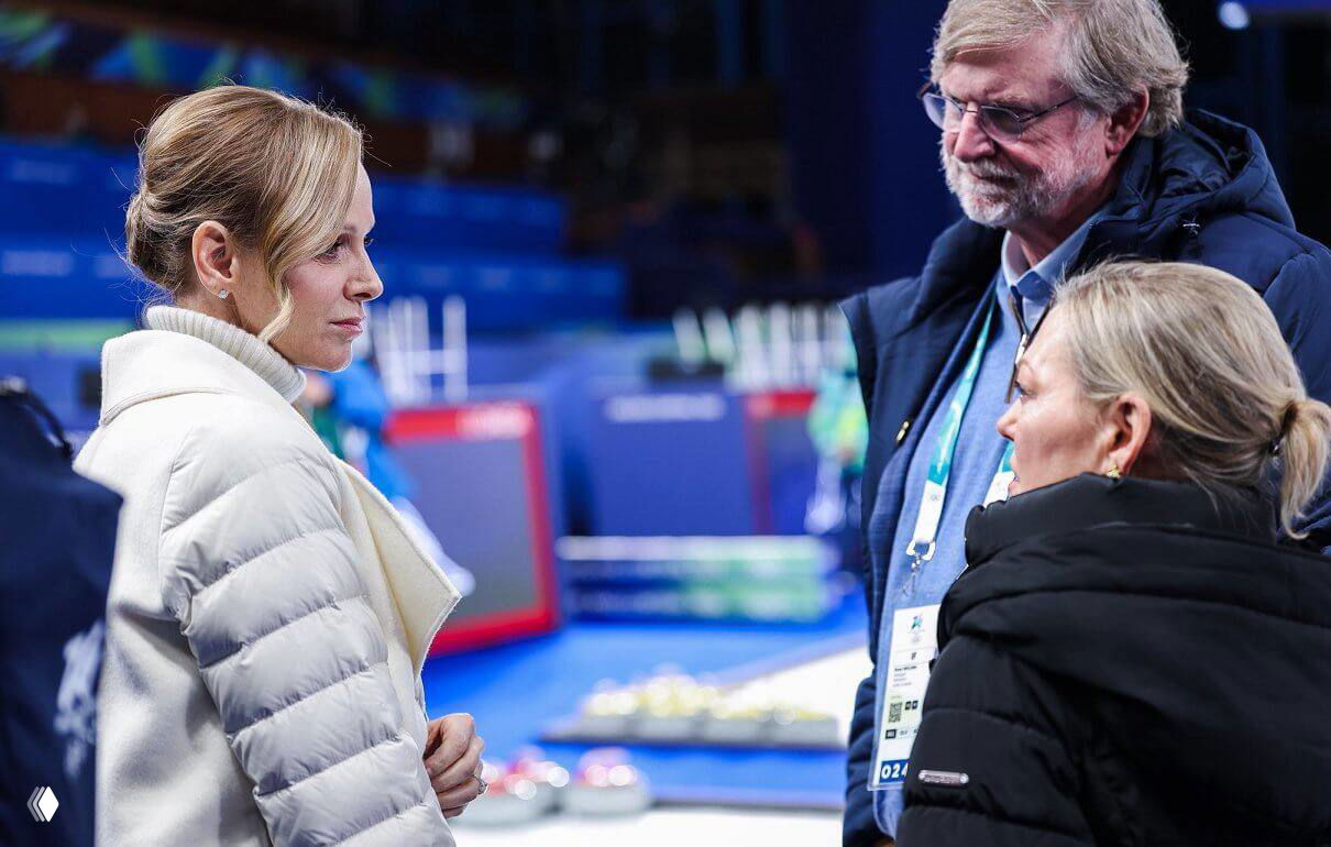 Княгиня Шарлен в белом пальто-пончо внимательно слушает организаторов у дорожки для керлинга, вокруг — официальные бэйджи и экипировка.