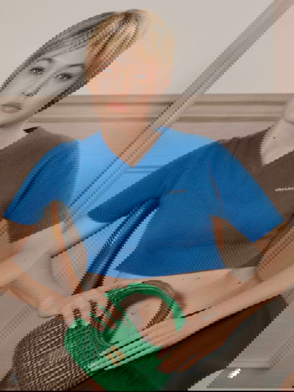 Модель с коротким париком в синем укороченном топе и зелёной сумкой на коленях — расслабленный кадр кампейна, подчёркивающий силуэт и текстуры.