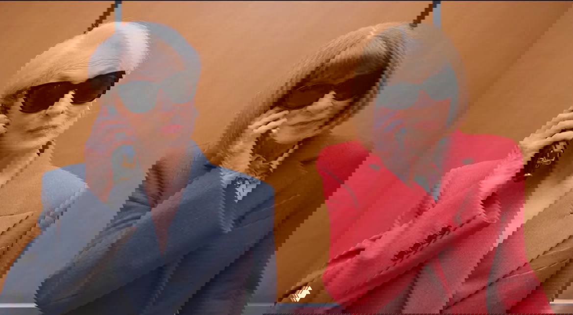 Крупный план в лифте: Анна Винтур и Мэрил Стрип разговаривают по телефону в строгих костюмах и тёмных очках — стилизованный кадр