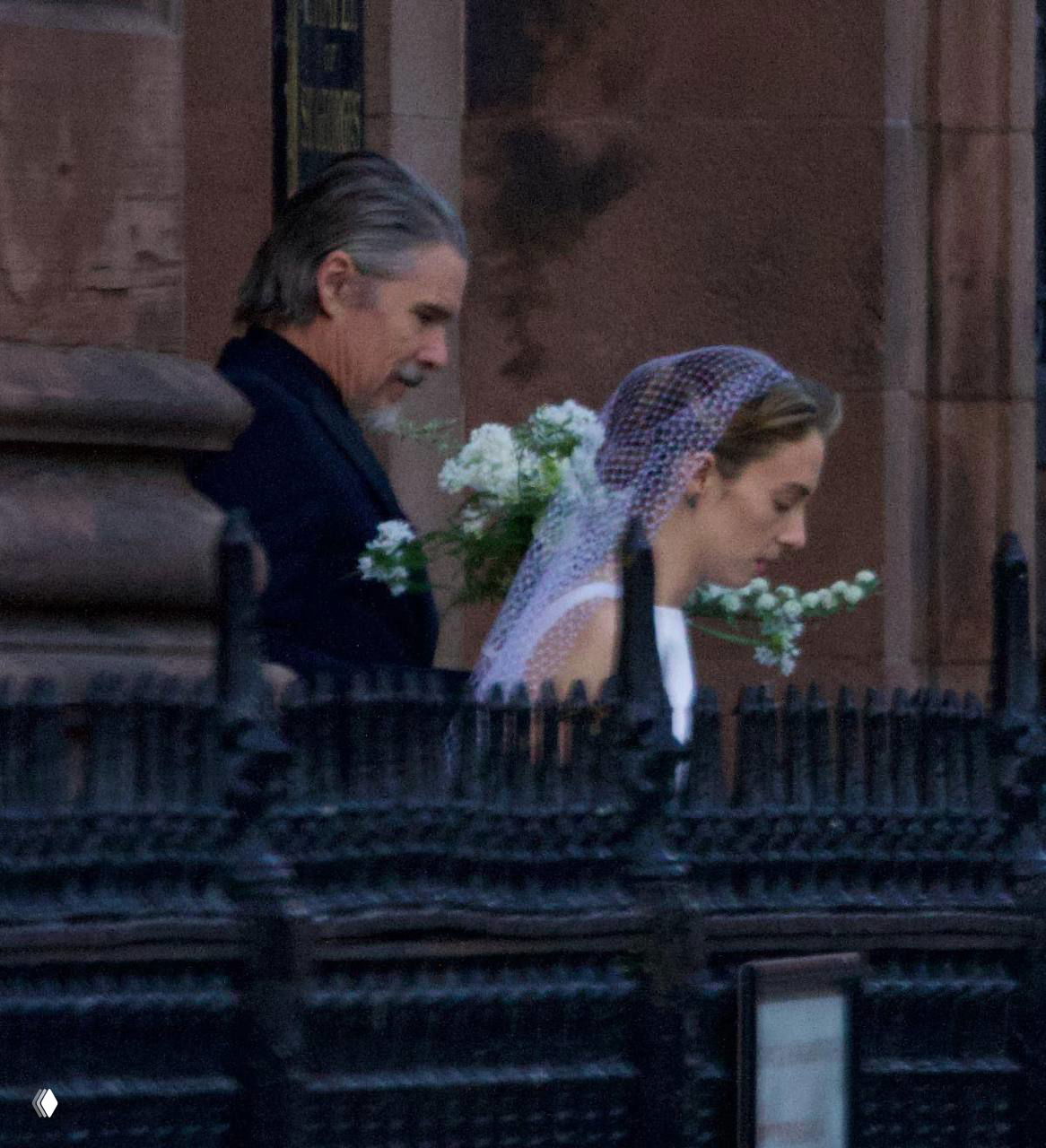 Крупный план: женщина в фате и мужчина рядом, они идут у каменной ограды церкви; видны букет и детали фаты.