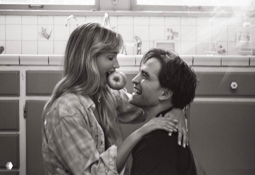 Игривый чёрно‑белый снимок: Лоуренс держит пончик у рта, Паттинсон улыбается — момент из той же фотосессии в домашней обстановке.