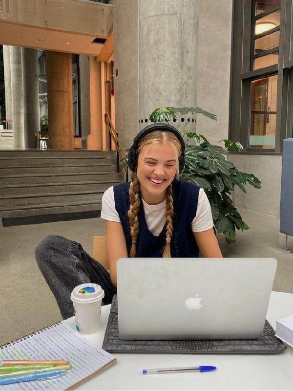 Улыбающаяся девушка в косичках и наушниках сидит за ноутбуком в общественном пространстве — учебная или рабочая обстановка.