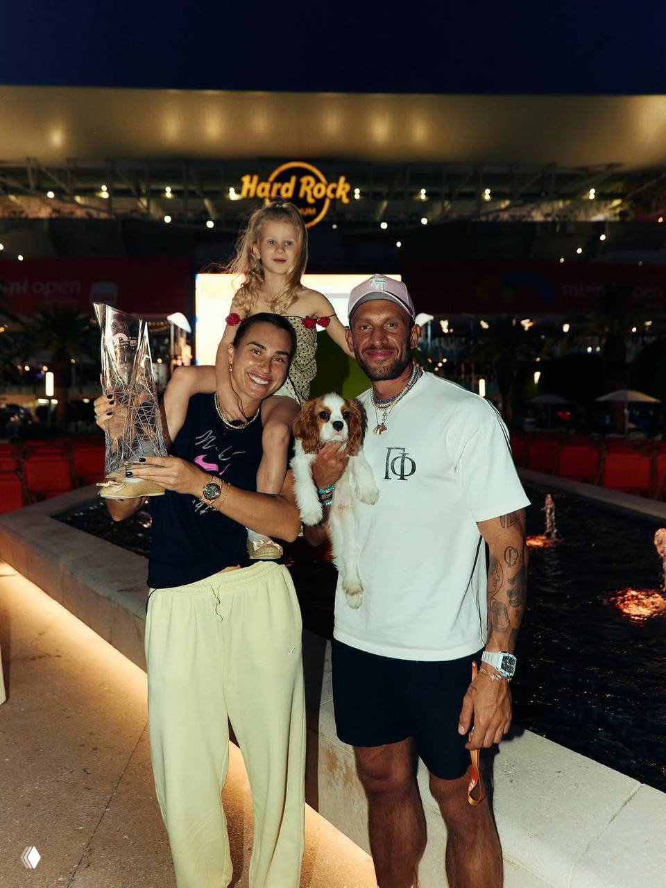 Групповое фото вечером у Hard Rock Stadium: Арина с трофеем, рядом семья, ребенок на плечах и маленькая собака на руках у одного из гостей.