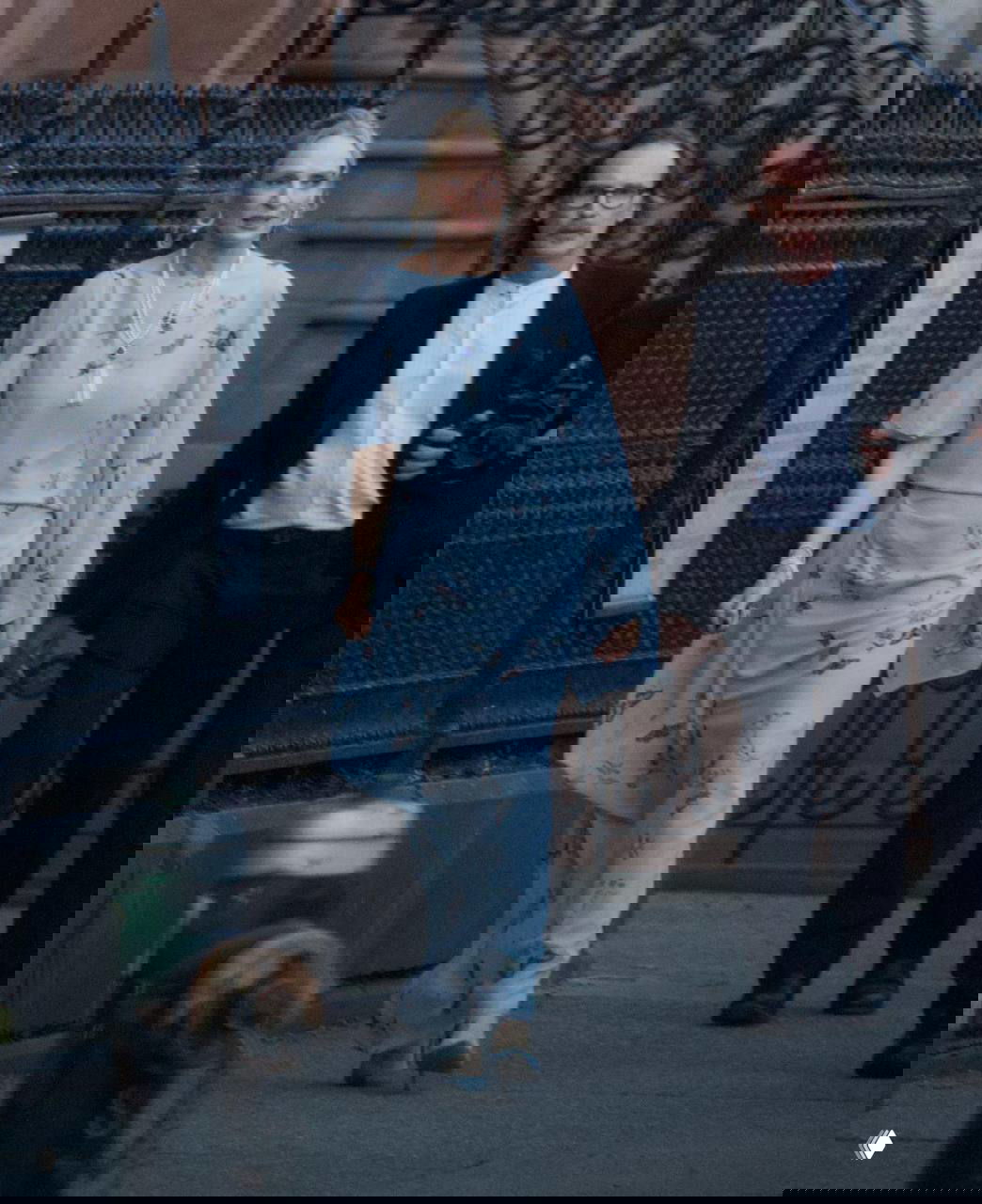 Гостья в светлом платье идёт по тротуару у ограды церкви; рядом мужчина с камерой сопровождает её на событии.