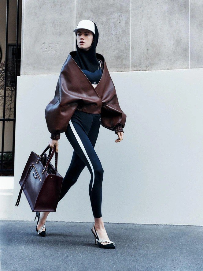 Модель в объёмной коричневой кожаной куртке и спортивных леггинсах несёт сумку и идёт по асфальту — спортивно-гламурный образ из коллекции.