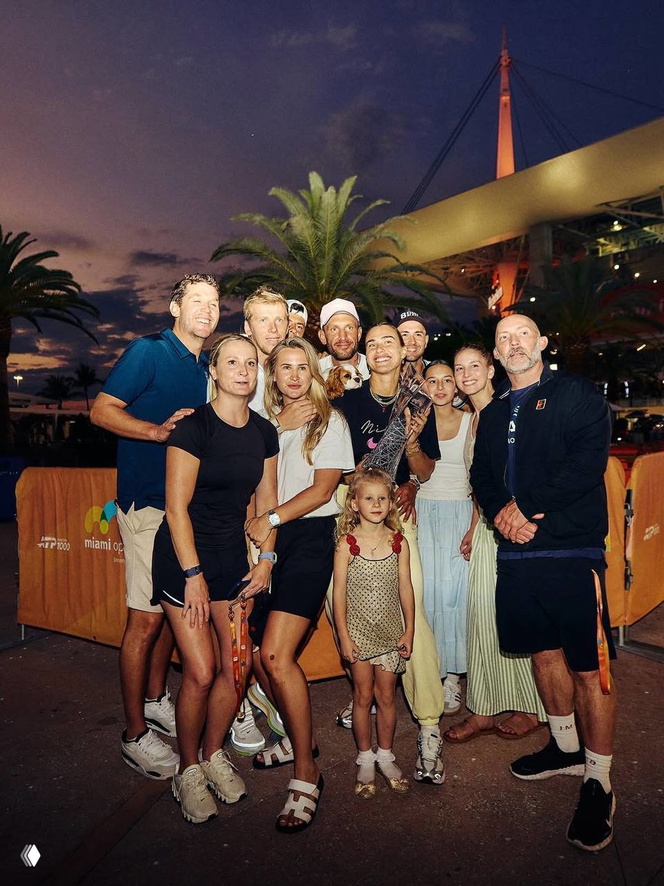 Большая совместная фотография команды и друзей Арины Соболенко на улице вечером: празднование победы, люди стоят группой на фоне освещённого неба.