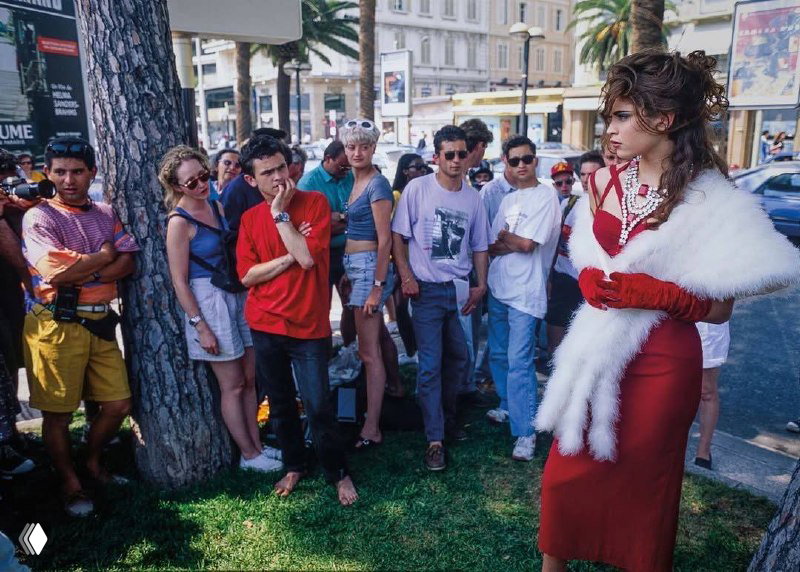 Уличная сцена Канн 1980‑х: модель в красном платье и белой накидке проходит мимо зрителей и фотографов, настроение ретро и глянца.