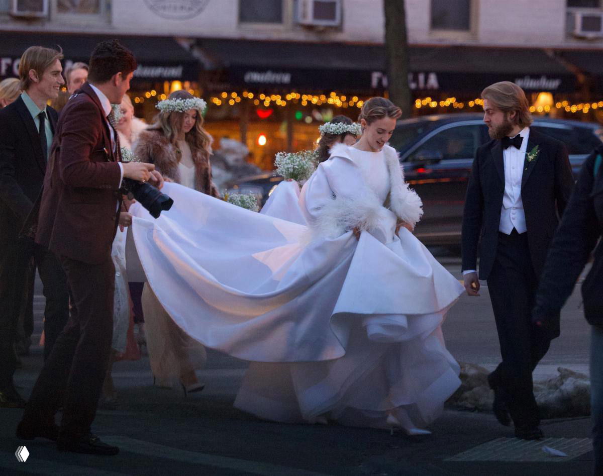 Невеста в белом платье с пышным подолом выходит на улицу, рядом жених в смокинге и гости с цветочными композициями.