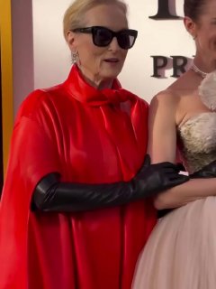 Кадр с близкого расстояния: женщина в красном плаще позирует вместе с партнёром и другими гостями на красной дорожке во время мировой премьеры фильма.