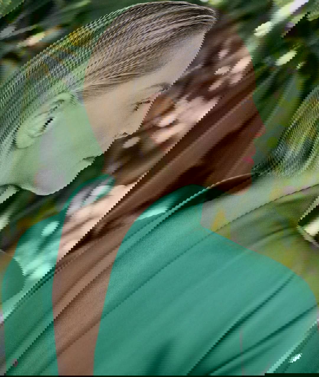 Профильный портрет женщины с короткой прической на фоне зелёной листвы — естественный макияж и спокойная поза в студийной атмосфере.