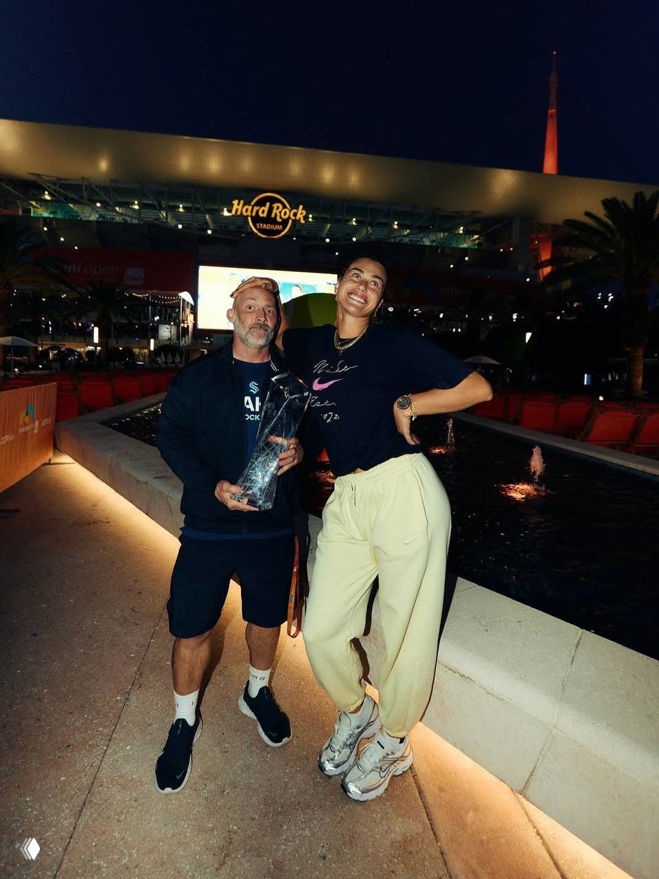 Ночная фотография на фоне входа в Hard Rock Stadium: Арина с трофеем позирует с мужчиной, в кадре вечерняя подсветка и фонтан позади.