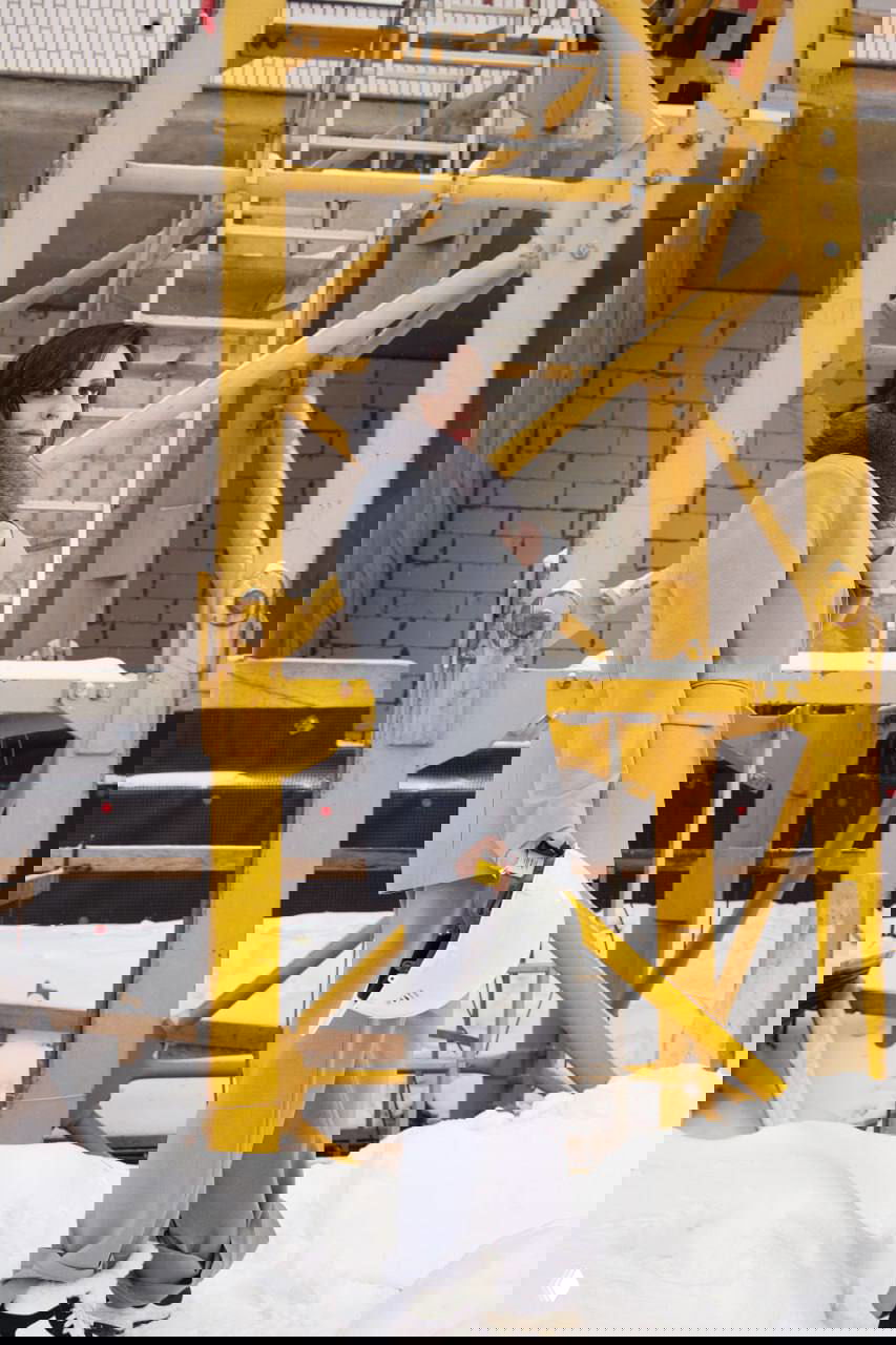 Женщина у жёлтой конструкции башенного крана в заснеённой строительной зоне, держит строительную каску в руке.