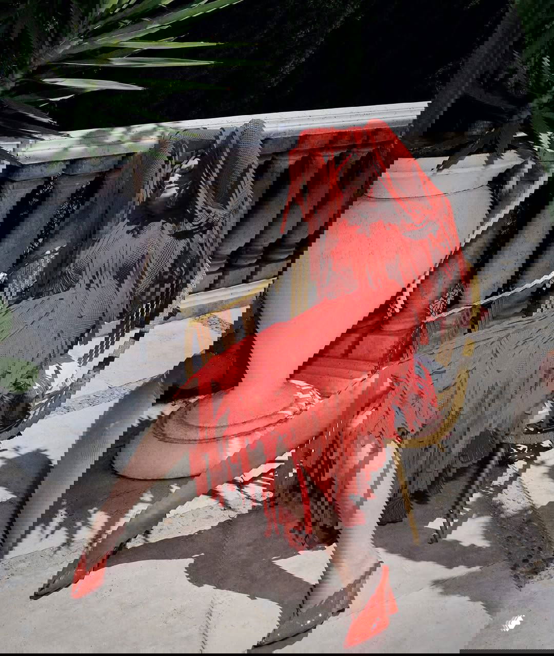 Женщина в ярком красном платье с бахромой расположилась в кресле на террасе, солнце подчёркивает текстуру ткани и тени вокруг.