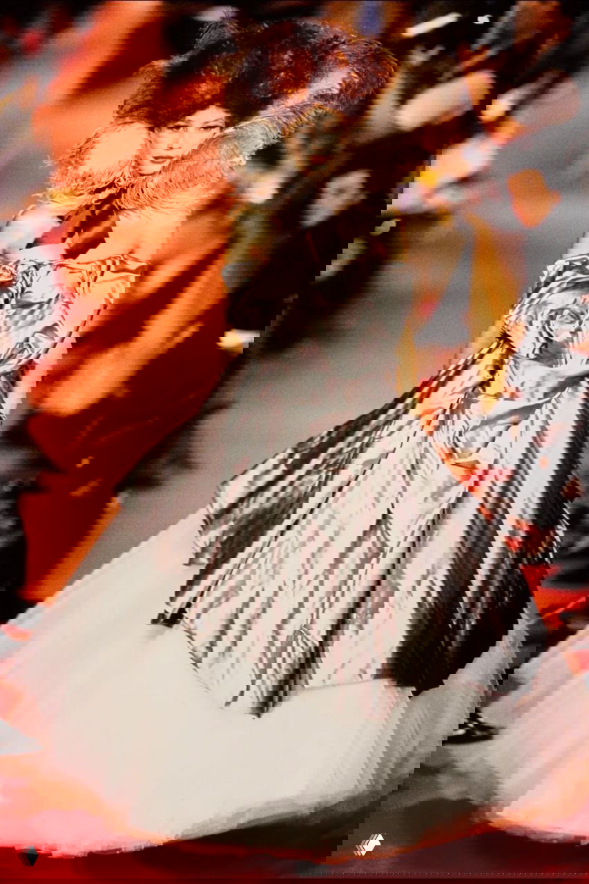 Модель в длинной тёмной шубе, сочетающей текстуры, несёт яркую сумку на показе Vivienne Westwood осень 1993 на красном подиуме.