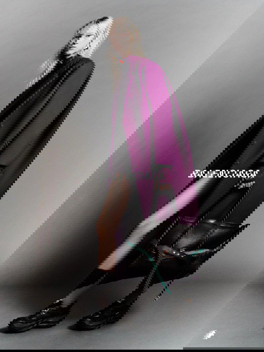 Высокая модель в длинном фиолетовом пальто и крупной сумке позирует на нейтральном фоне — рекламный кадр с логотипом Balenciaga.